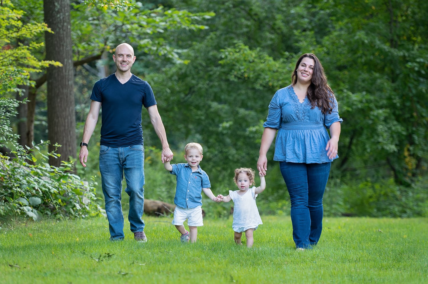 Family of four in denim walking hand in hand across the summer lawn at Bartlett Park in Chelmsford Massachusetts