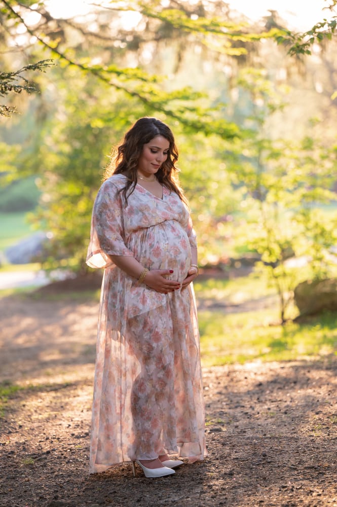 Expecting mother standing on the shaded path in a long floral dress at Benson Park in Hudson NH