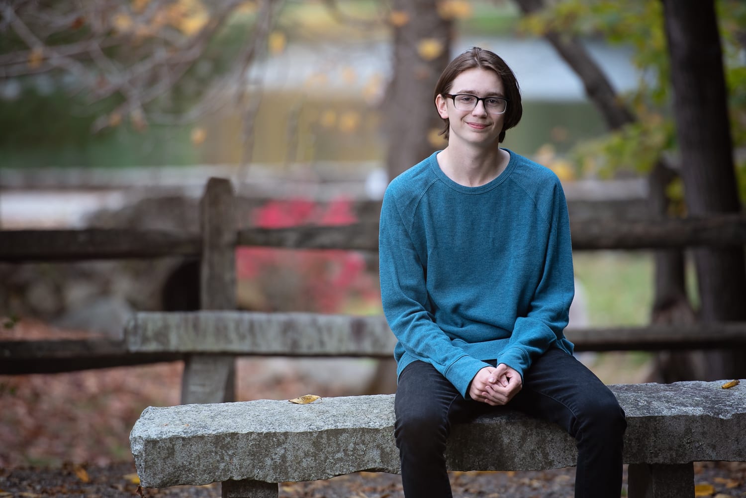 High school senior seated on a granite bench with a rustic fence and autumn color behind at Benson Park, Hudson NH