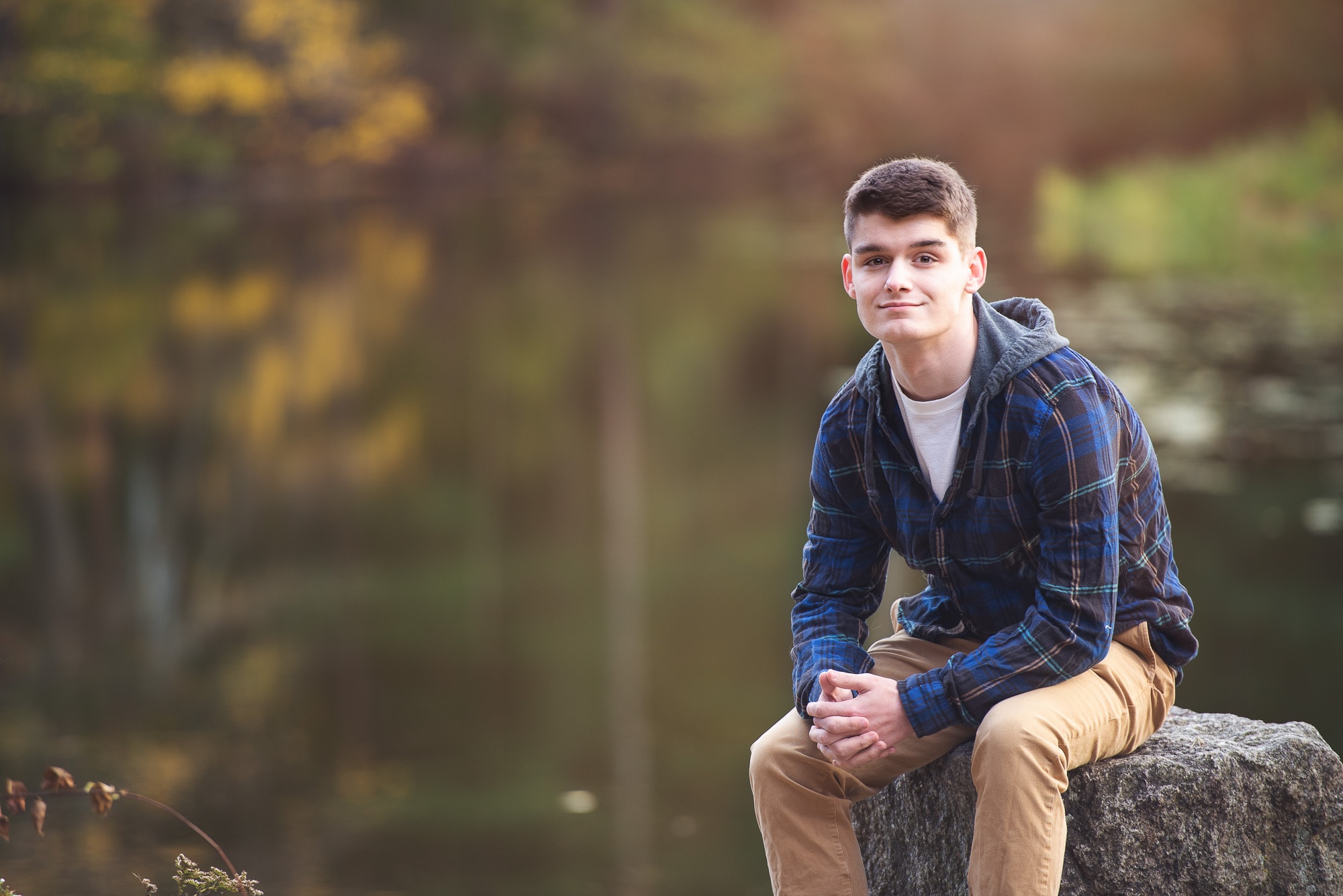 High school senior seated on a rock at the edge of Meadow Pond in autumn at Great Brook Farm State Park in Carlisle MA