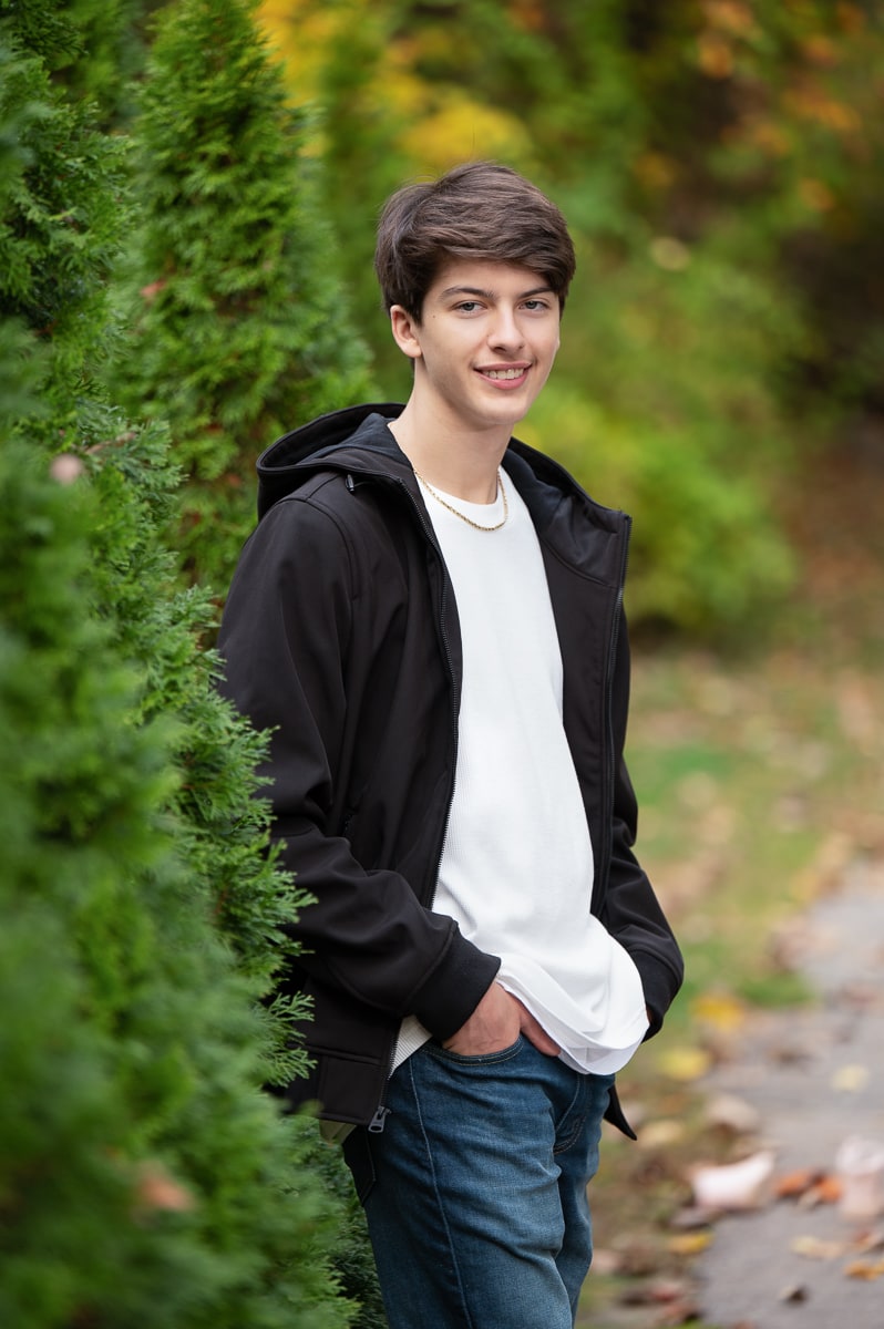 High school senior standing against a tall evergreen hedge at Benson Park, Hudson New Hampshire