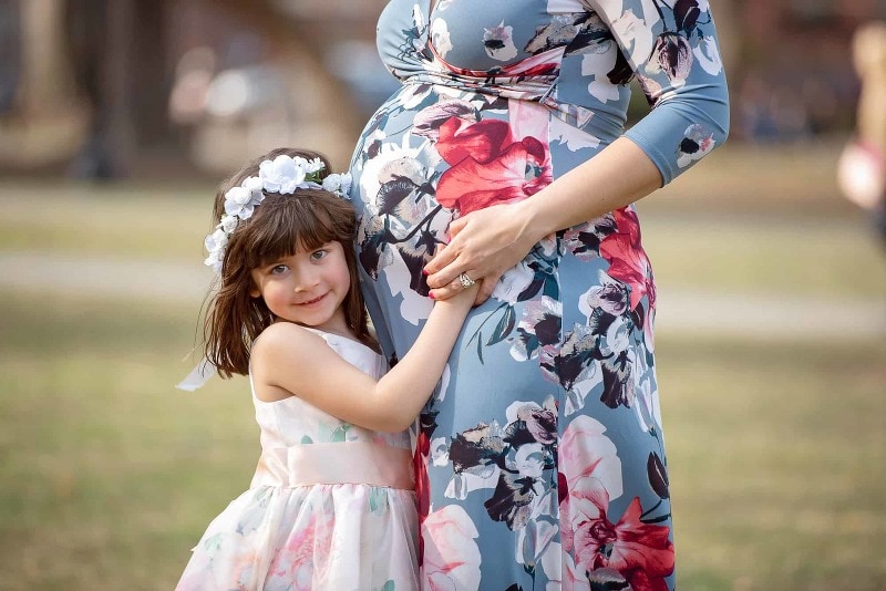 Young girl in a white dress and flower crown holding her expecting mother's baby bump on the lawn in the Boston Public Garden in Boston MA