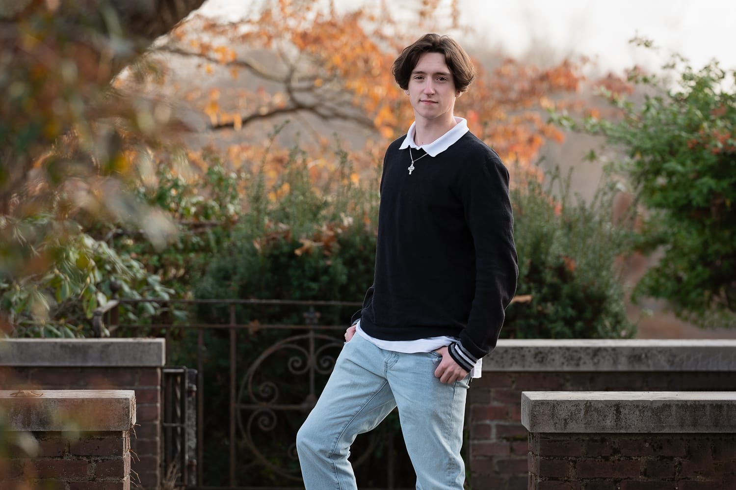 High school senior boy leaning against a brick wall by an iron gate in autumn in Concord MA