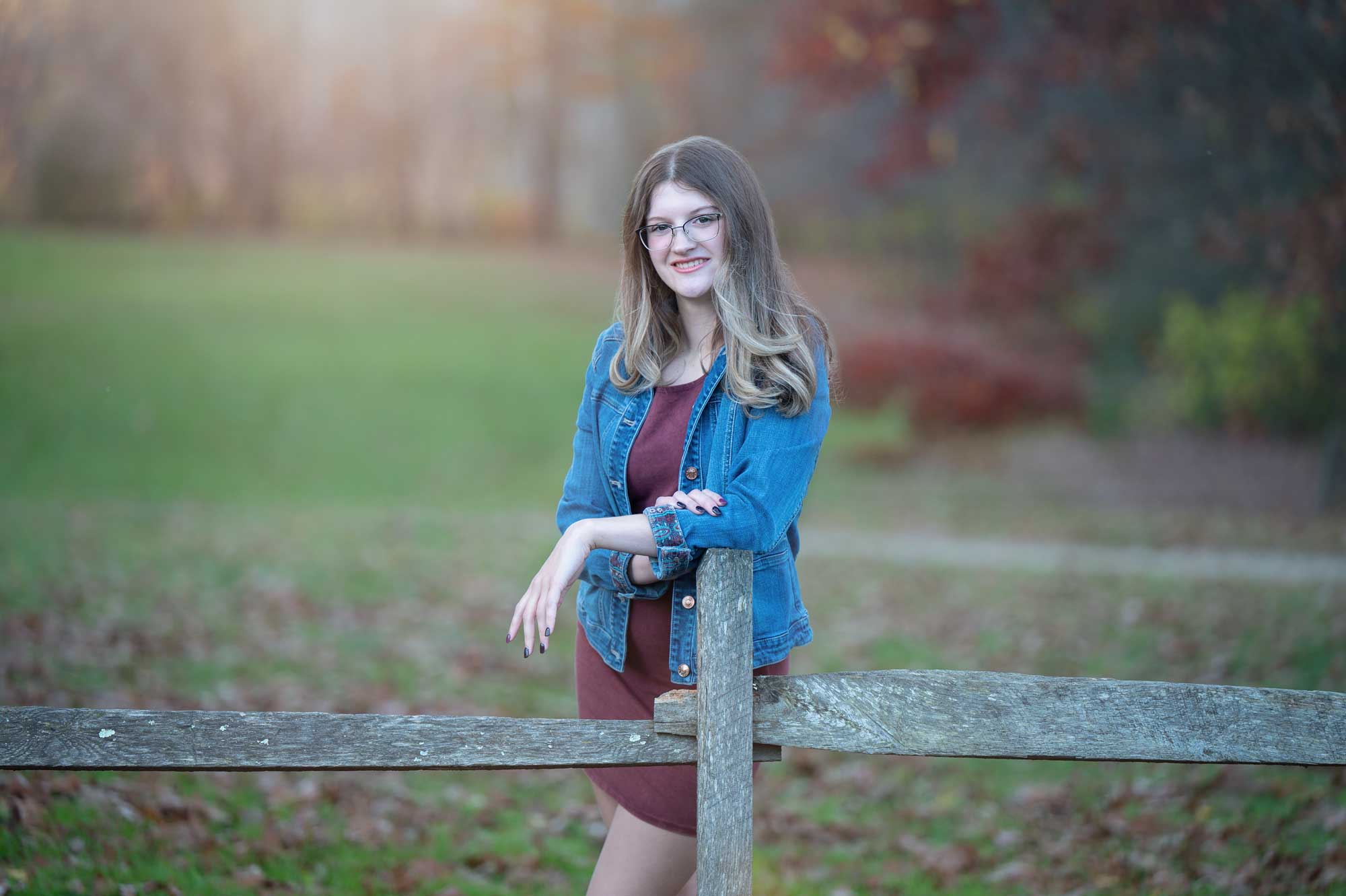 High school senior girl in a denim jacket at a split-rail fence in autumn at Great Brook Farm in Carlisle MA