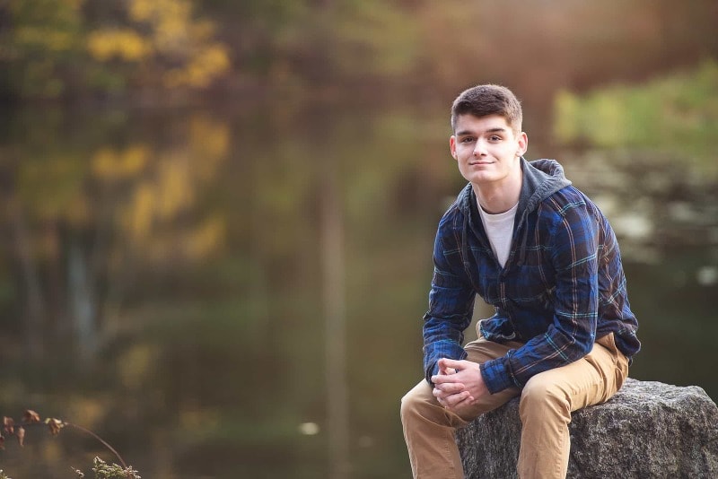 senior portrait in the fall by a pond sitting on a rock at great brook farm park in carlisle, ma