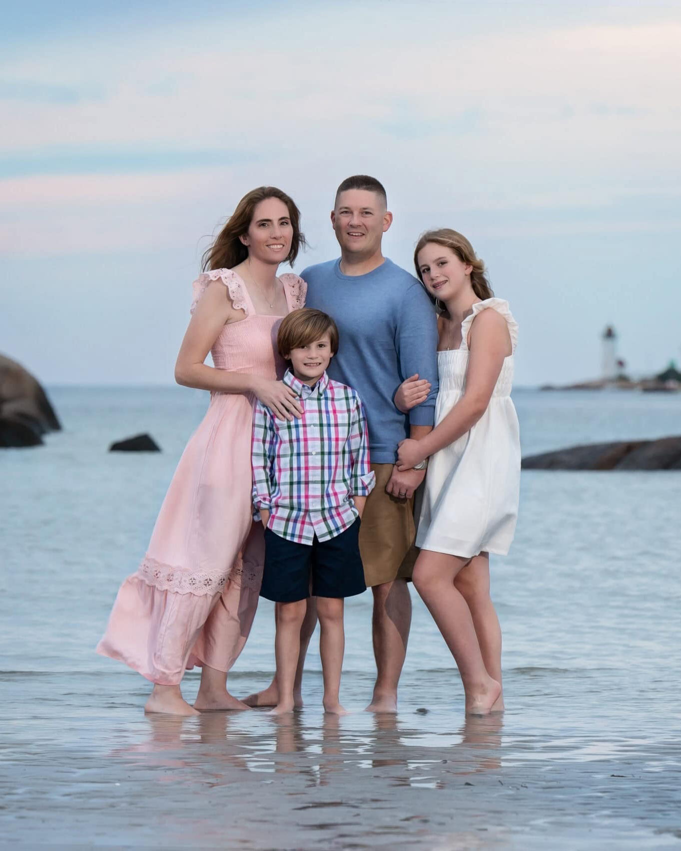 Family of four standing in the shallow water at Wingaersheek Beach in Gloucester MA with Annisquam Harbor Light behind them