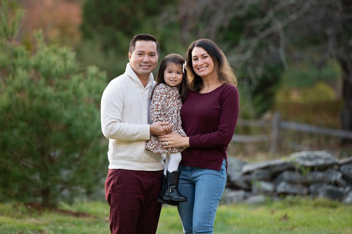 Parents holding their young daughter beside a stone wall by the Concord River in autumn in Concord MA
