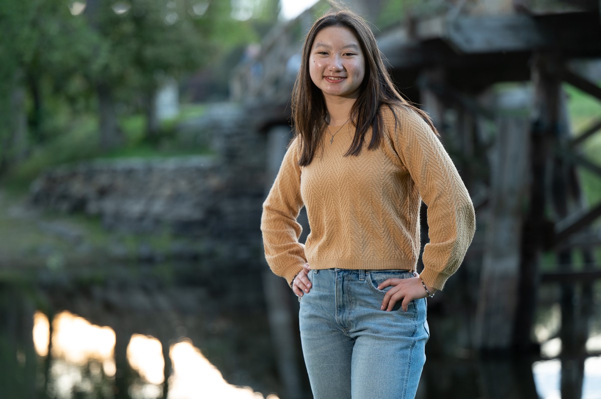 High school senior girl standing by the river with a wooden bridge behind her in Concord MA
