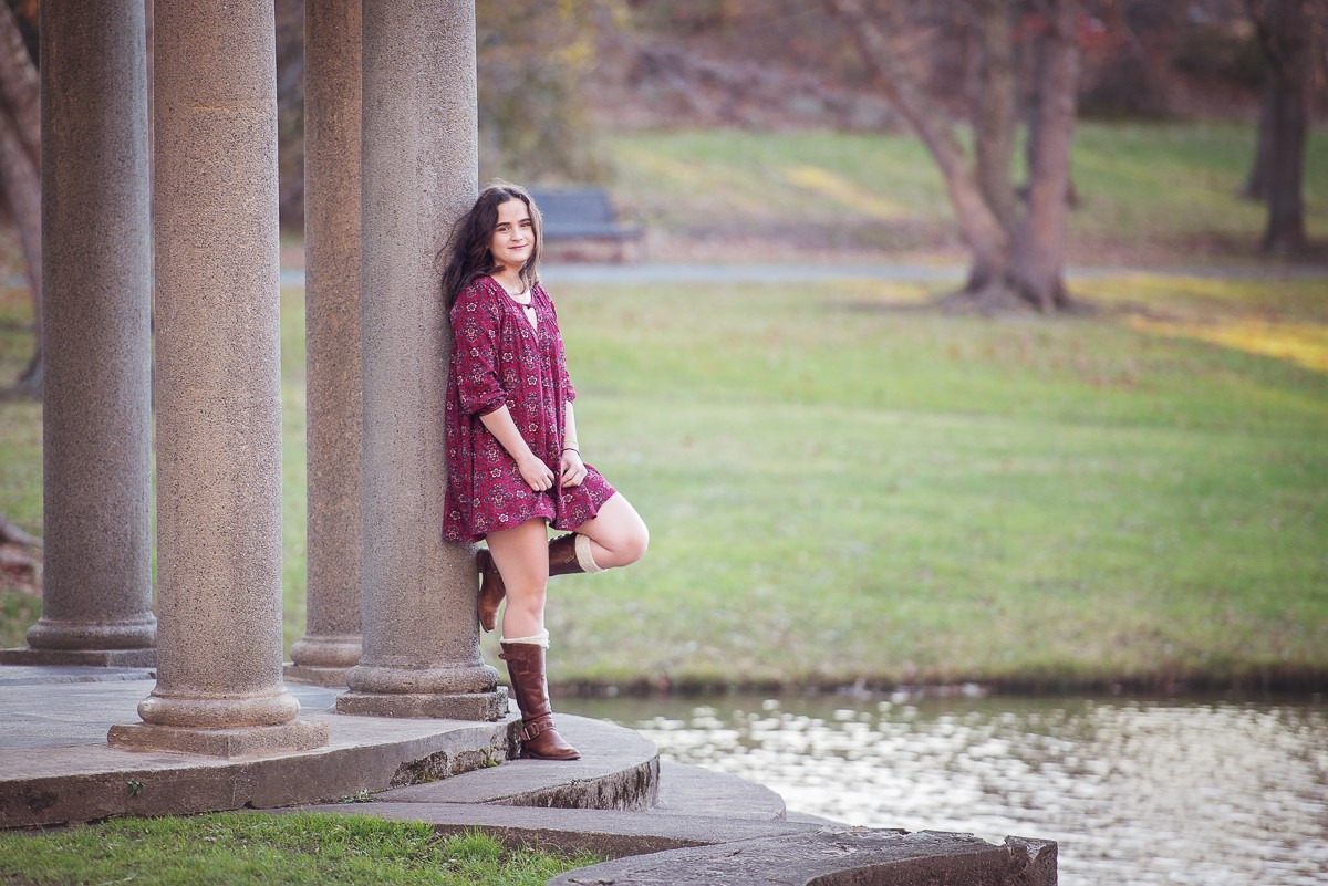 High school senior in a burgundy floral dress leaning against a stone column above the lagoon at Larz Anderson Park in Brookline MA