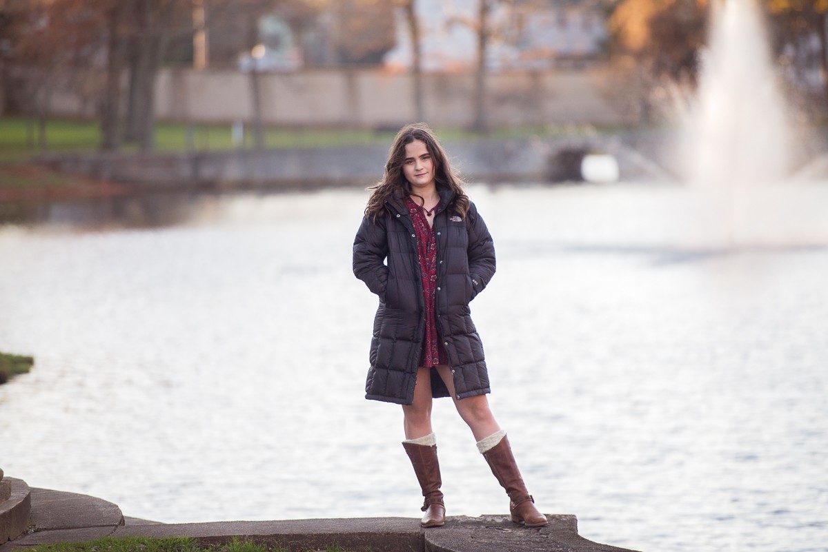 High school senior in a winter coat standing on the stone landing at the lagoon with the fountain behind her at Larz Anderson Park in Brookline Massachusetts