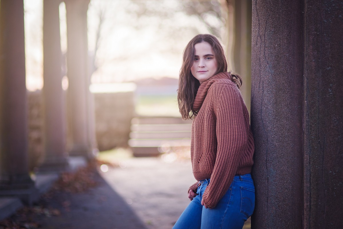 High school senior in a rust sweater leaning against a stone column with the low sun behind her at Larz Anderson Park in Brookline MA