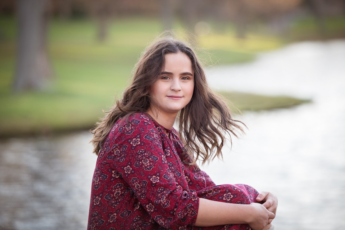 High school senior seated at the edge of the lagoon in a burgundy floral dress at Larz Anderson Park in Brookline Massachusetts