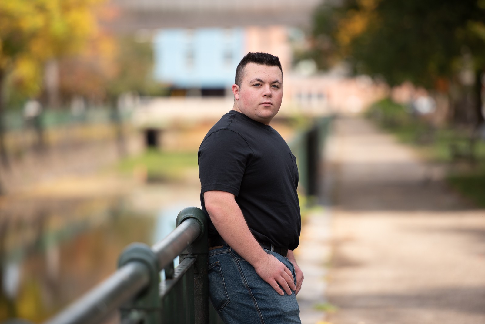 High school senior leaning on the iron rail of the canal walkway with the towpath and brick buildings behind him in Lowell MA, photographed by Chelmsford portrait photographer Luciana Calvin