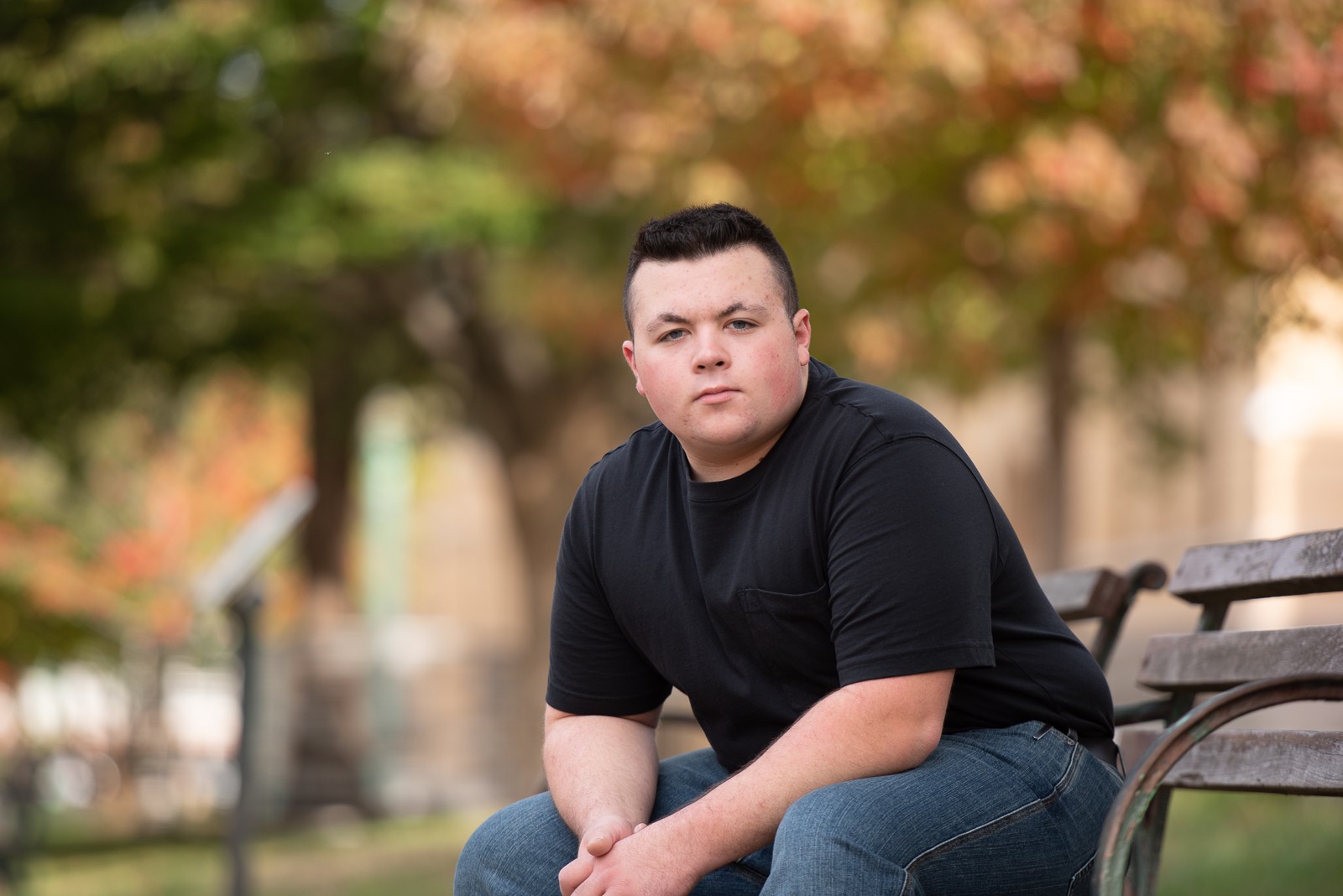 High school senior seated on a bench under autumn maples with a brick mill wall behind him in Lowell MA