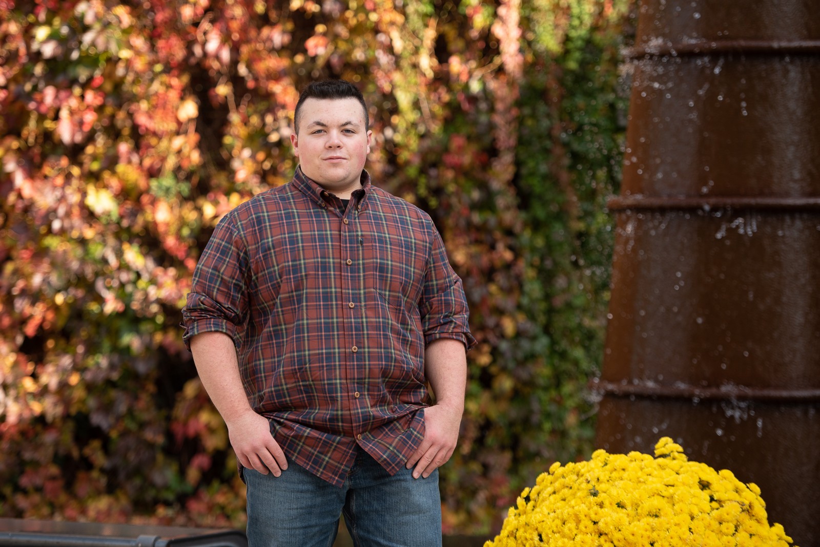 High school senior in a plaid shirt standing in front of a wall of red autumn Boston ivy beside a copper fountain and yellow mums in Lowell MA