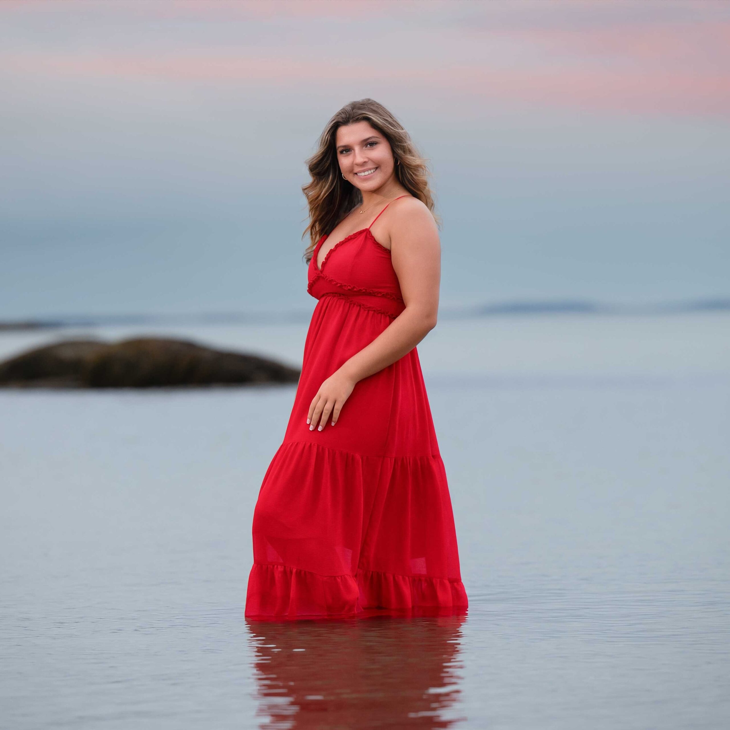 High school senior girl in a red gown standing in the shallow water at Wingaersheek Beach in Gloucester MA