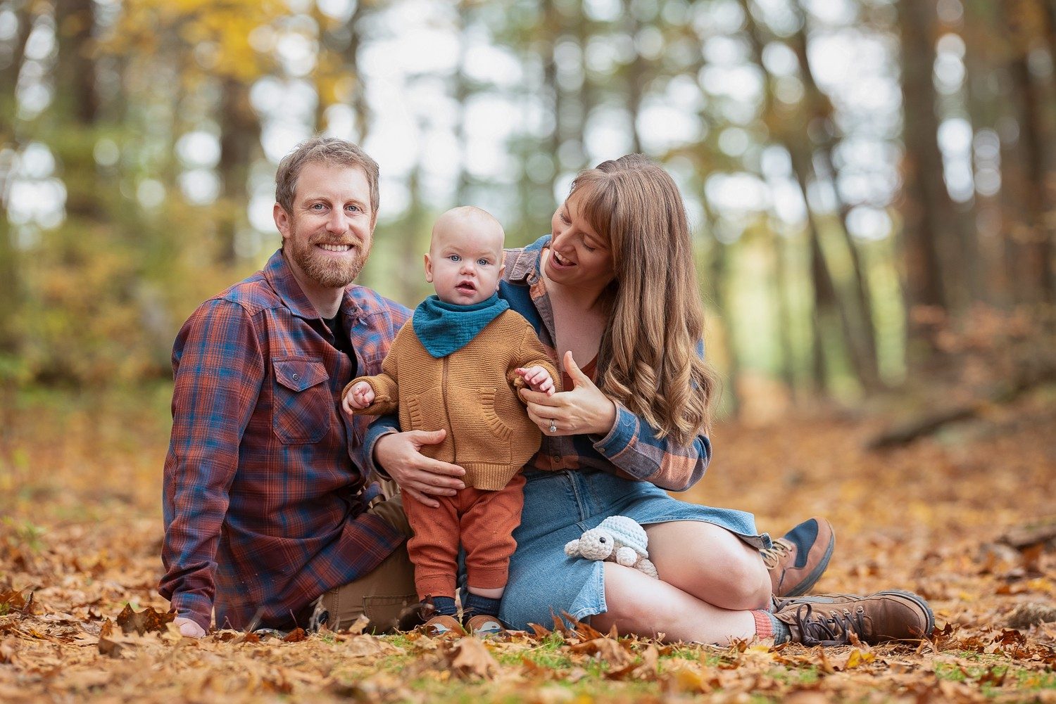 Parents kneeling in the leaf fall with their baby between them at Assabet River National Wildlife Refuge in Sudbury Massachusetts