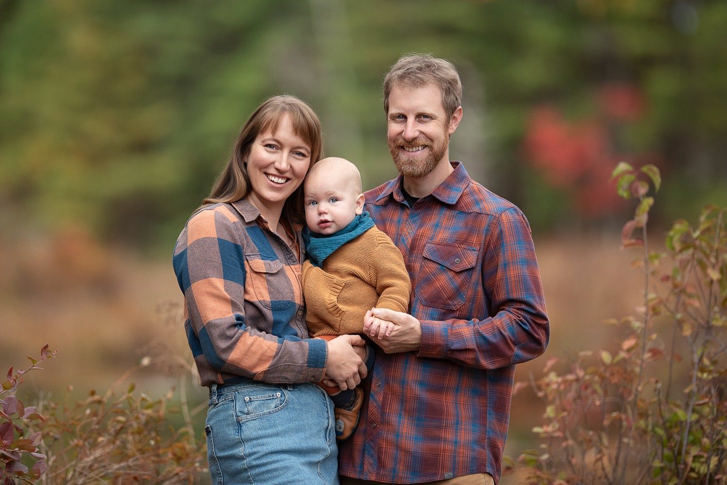 Parents holding their baby between them with red autumn foliage behind at Assabet River National Wildlife Refuge in Sudbury MA