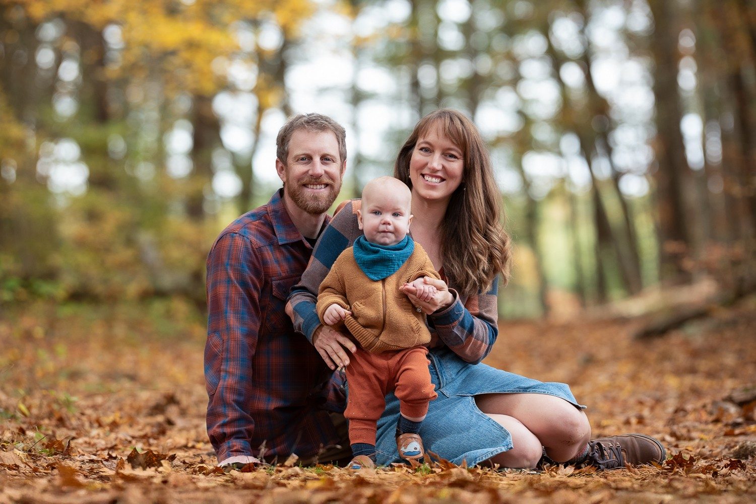 Family of three sitting together in deep fallen leaves under tall pines at Assabet River National Wildlife Refuge, Sudbury MA