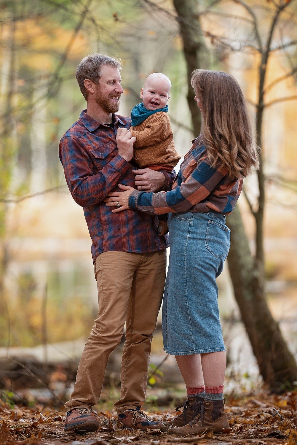 Parents facing each other holding their baby between them beside a slender tree at Assabet River National Wildlife Refuge in Sudbury MA