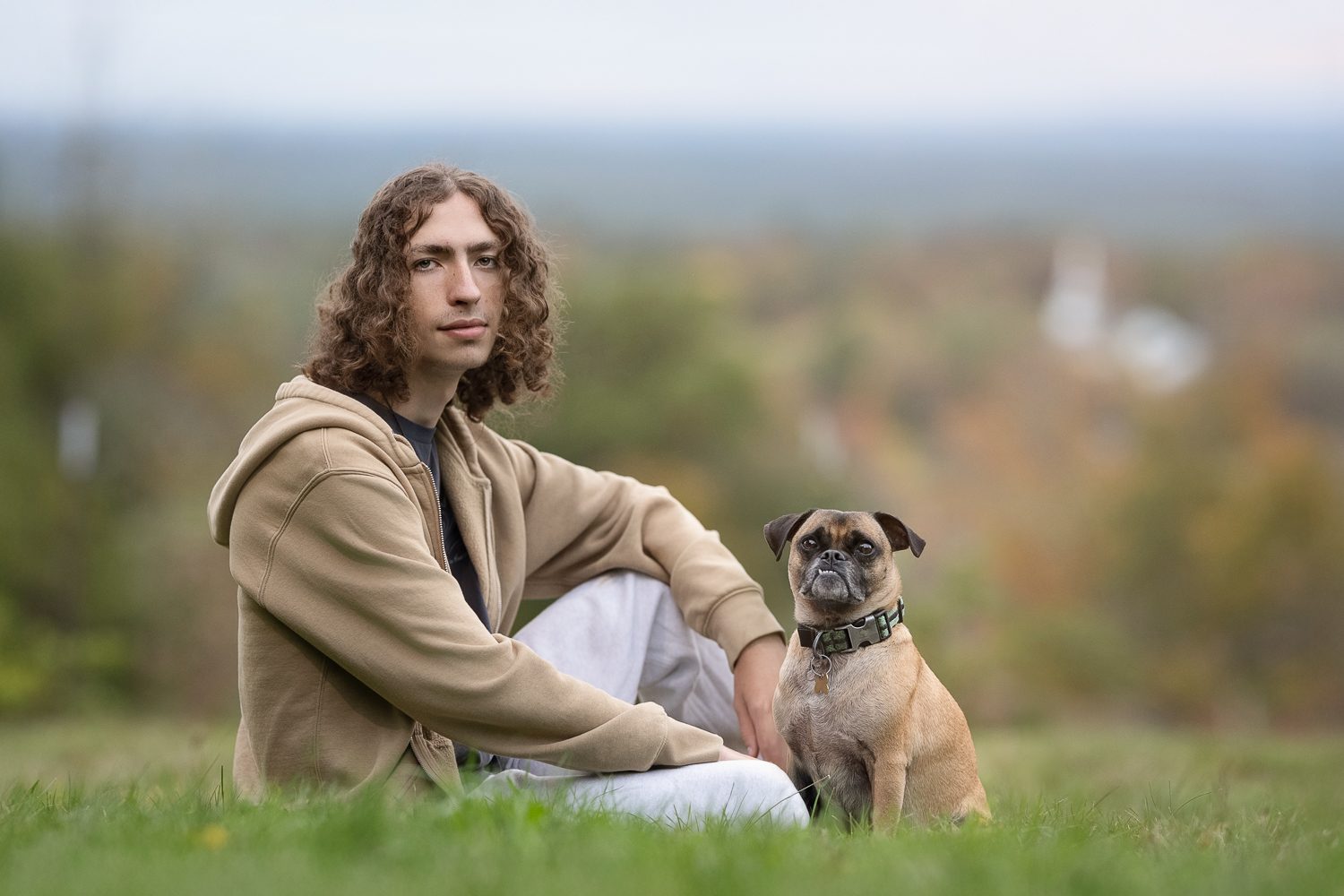 High school senior sitting on the summit grass with his dog and the Nashoba Valley behind him at Gibbet Hill in Groton MA