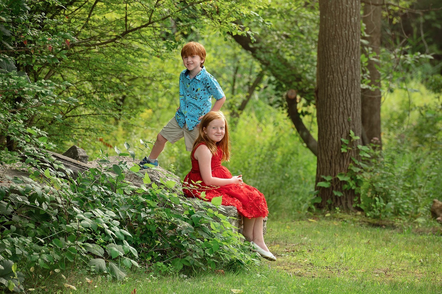 Young boy and his sister in a red dress sitting on the rocks among green summer woods at Bartlett Park in Chelmsford MA