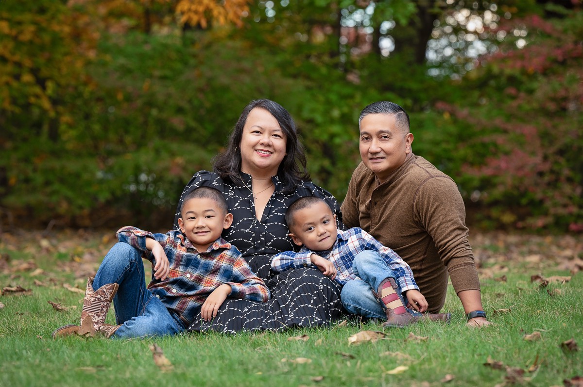 Family of four sitting together on the lawn with autumn color behind them at Bartlett Park in Chelmsford MA