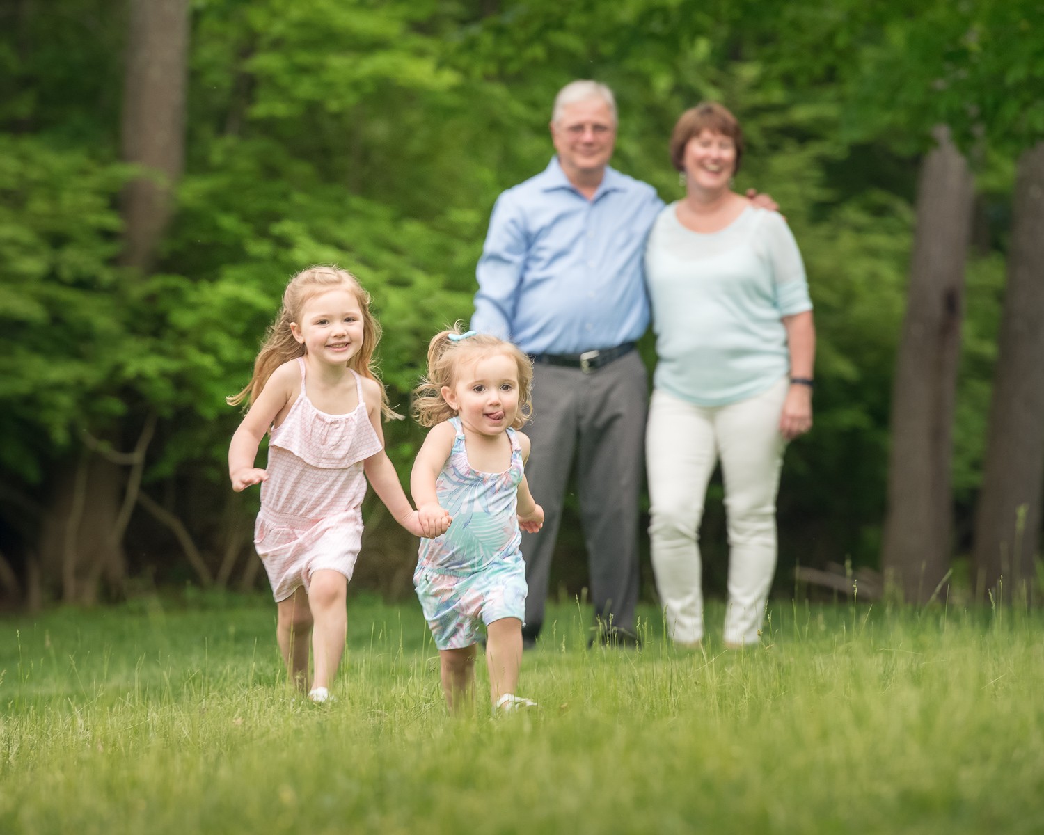 Two little girls in pastel dresses running across the lawn toward the camera with their grandparents behind them at Bartlett Park in Chelmsford MA