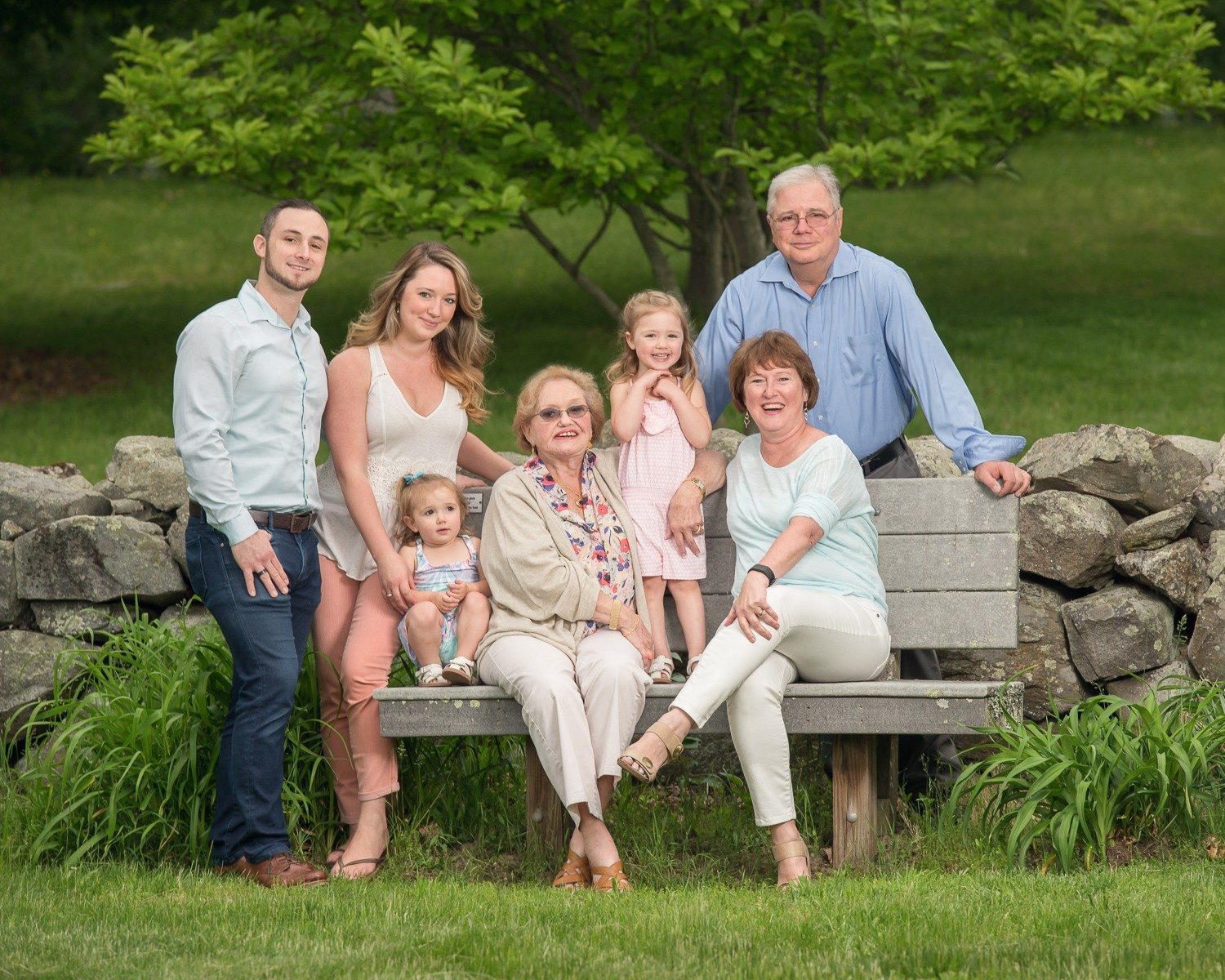 Extended family of eight gathered on and around a wooden bench with a fieldstone wall behind them at Bartlett Park in Chelmsford Massachusetts