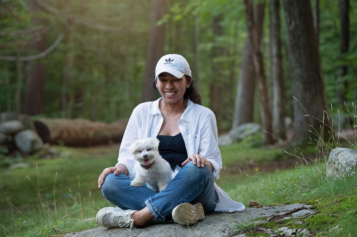 Woman seated on a rock holding her small white dog in the green woods at Bartlett Park in Chelmsford Massachusetts