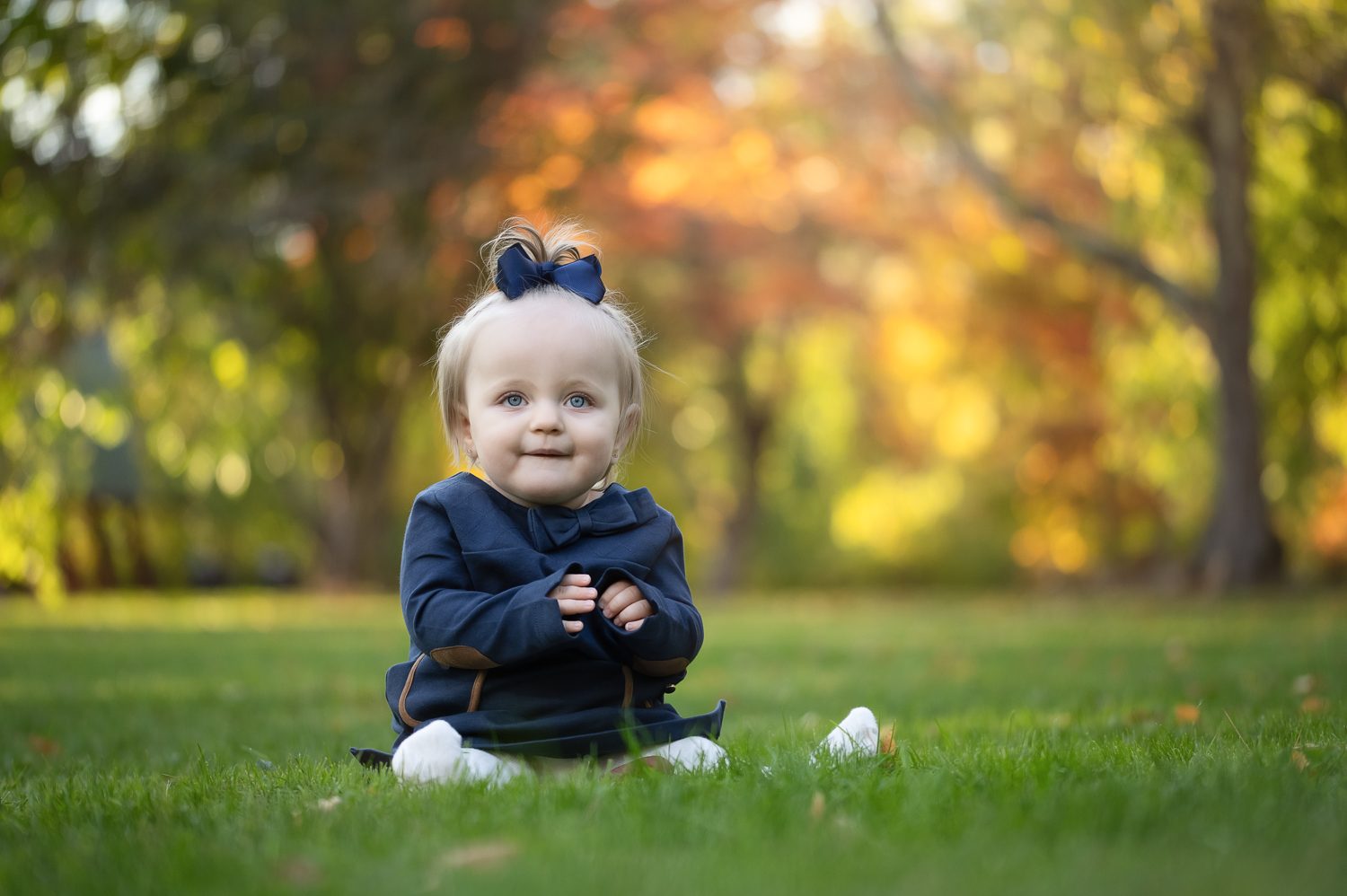 Baby in navy sitting alone on the lawn with golden autumn trees behind at Benson Park in Hudson NH