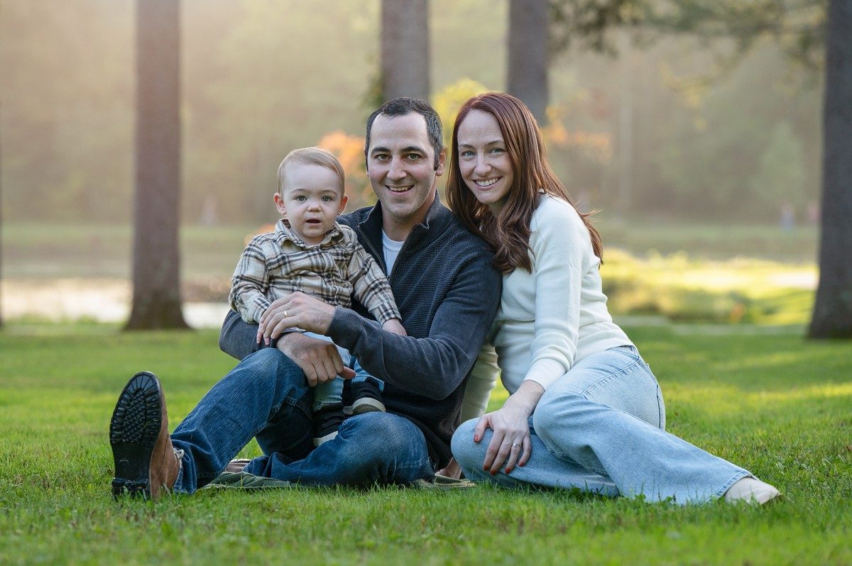 Young family of three sitting on the lawn in golden evening light at Benson Park, Hudson NH