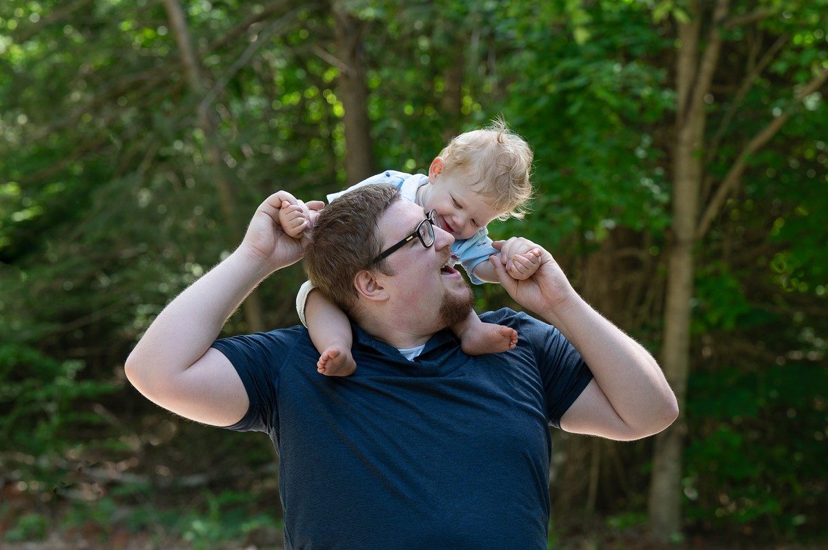 Father carrying his toddler on his shoulders under summer trees at Benson Park, Hudson New Hampshire