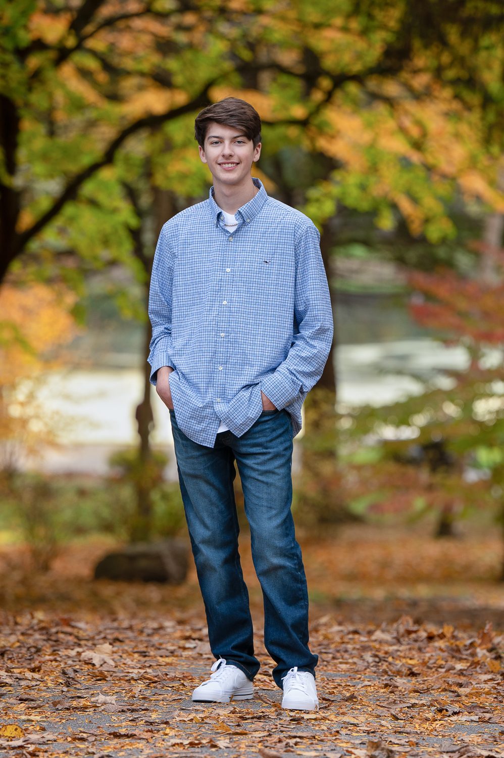 High school senior standing on the lawn with the pond and autumn trees behind him at Benson Park in Hudson NH