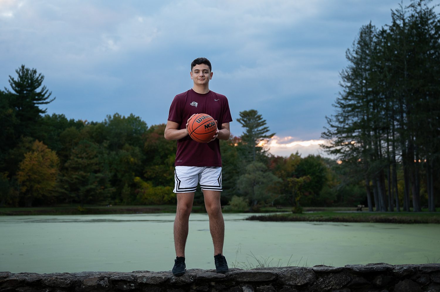 High school senior standing on the stone wall with a basketball and the pond and evening sky behind him at Benson Park in Hudson NH