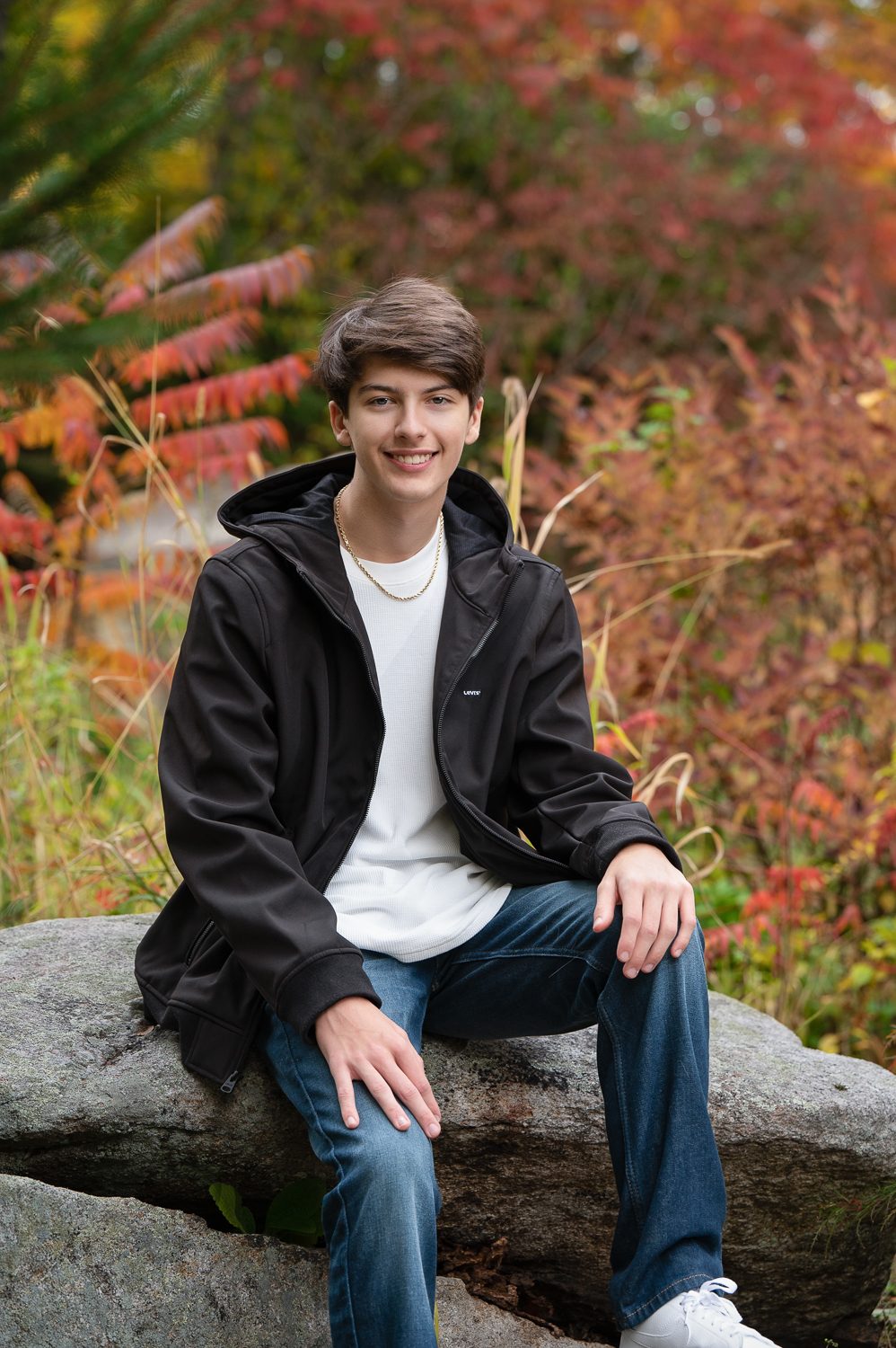 High school senior seated on a glacial boulder with a red maple behind him at Benson Park in Hudson NH