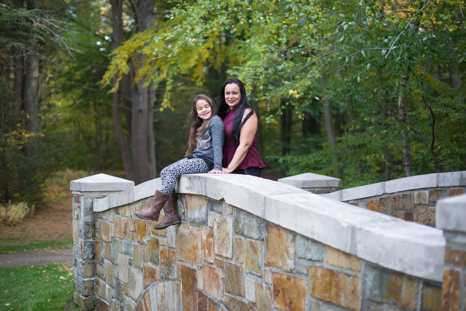 Mother and her daughter sitting on the parapet of an artisan-built stone bridge at Bird Park in Walpole MA with autumn trees behind them