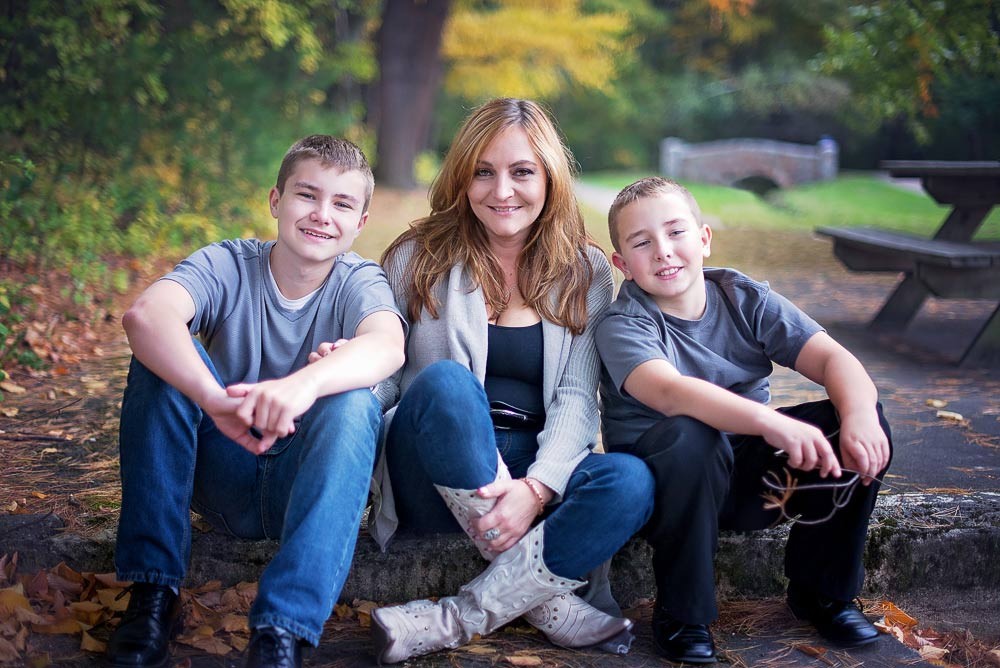 Mother seated on a stone step with her two sons among fallen leaves at Bird Park in Walpole Massachusetts, a stone bridge behind them