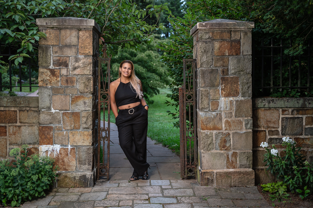 High school senior in black standing in the cut stone gateway with wrought iron gates at Francis William Bird Park in Walpole MA