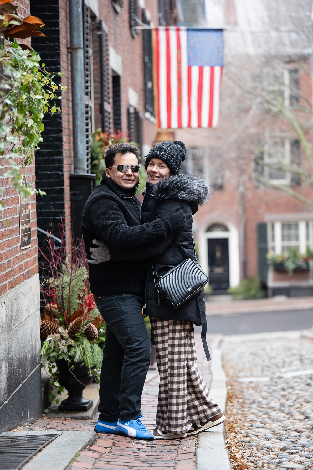 Couple in winter coats on the cobblestones of Acorn Street in Beacon Hill Boston with an American flag above them