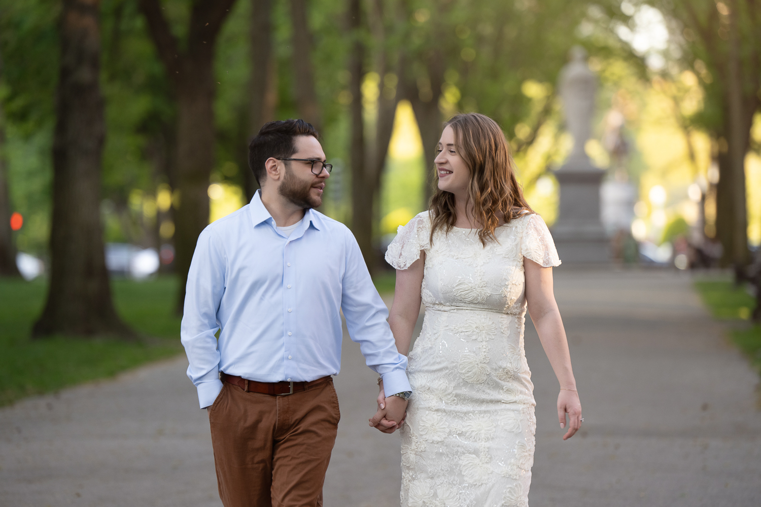 Engaged couple walking hand in hand down the tree-lined Commonwealth Avenue Mall with a statue behind them in Boston Massachusetts