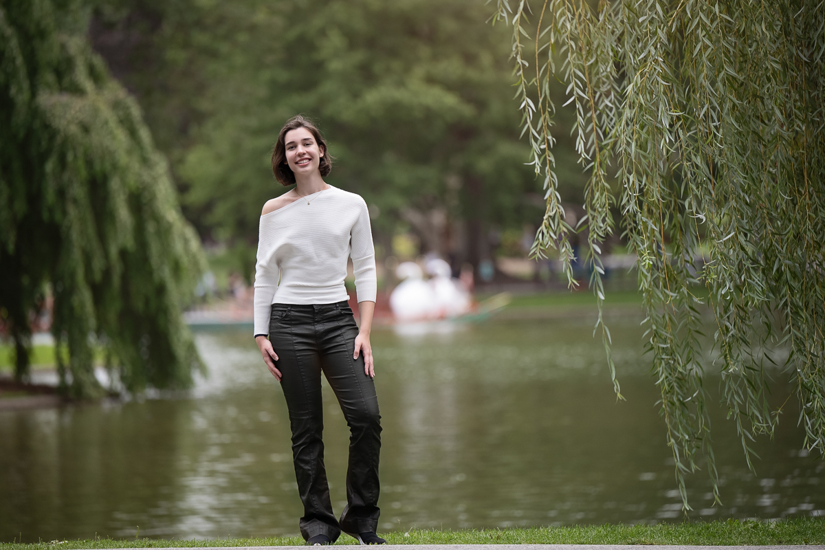 High school senior standing under the weeping willows by the lagoon with a Swan Boat passing behind her in the Boston Public Garden