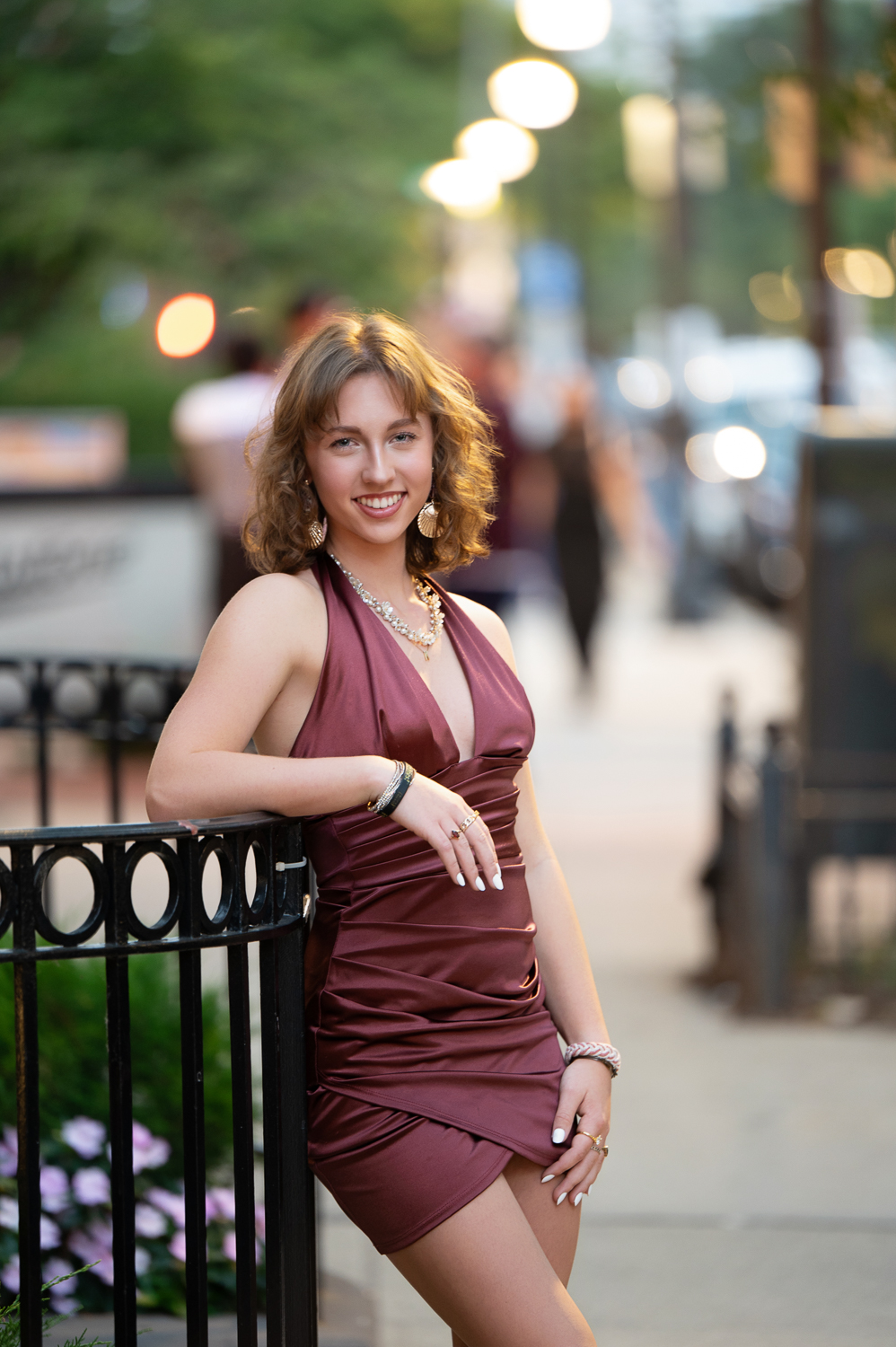 High school senior in a burgundy dress leaning on a wrought iron fence on Newbury Street in Boston MA with the shopfront lights coming on behind her