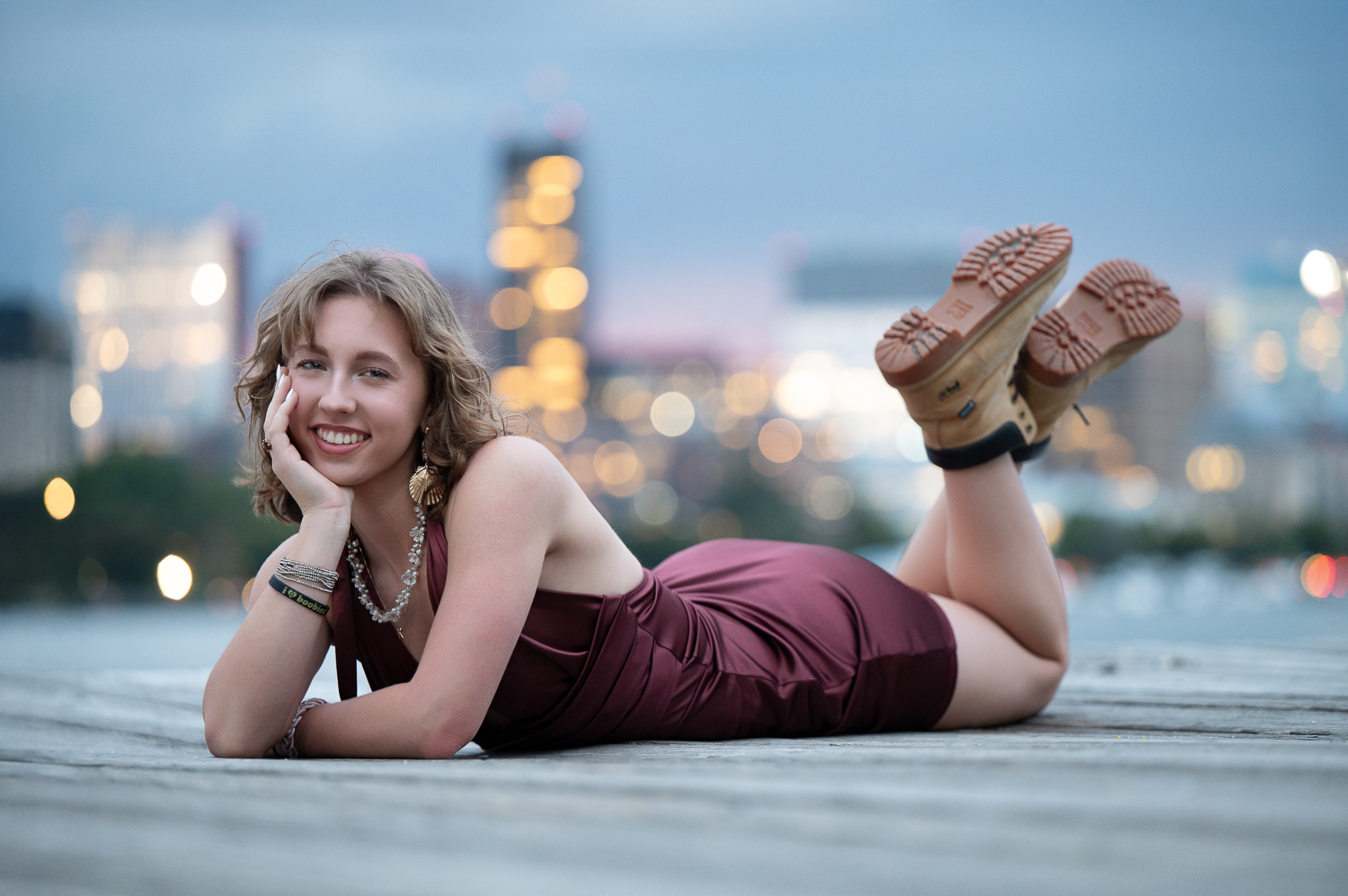 High school senior lying on a boardwalk at blue hour with the Boston skyline lit behind her in Boston Massachusetts