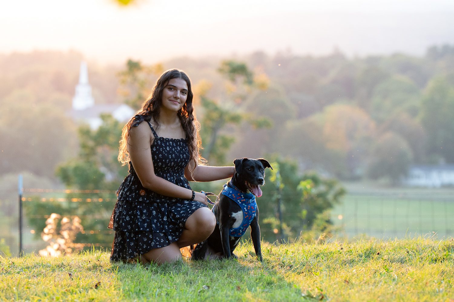 High school senior kneeling on the hilltop grass with her black dog and the valley behind at Gibbet Hill in Groton Massachusetts