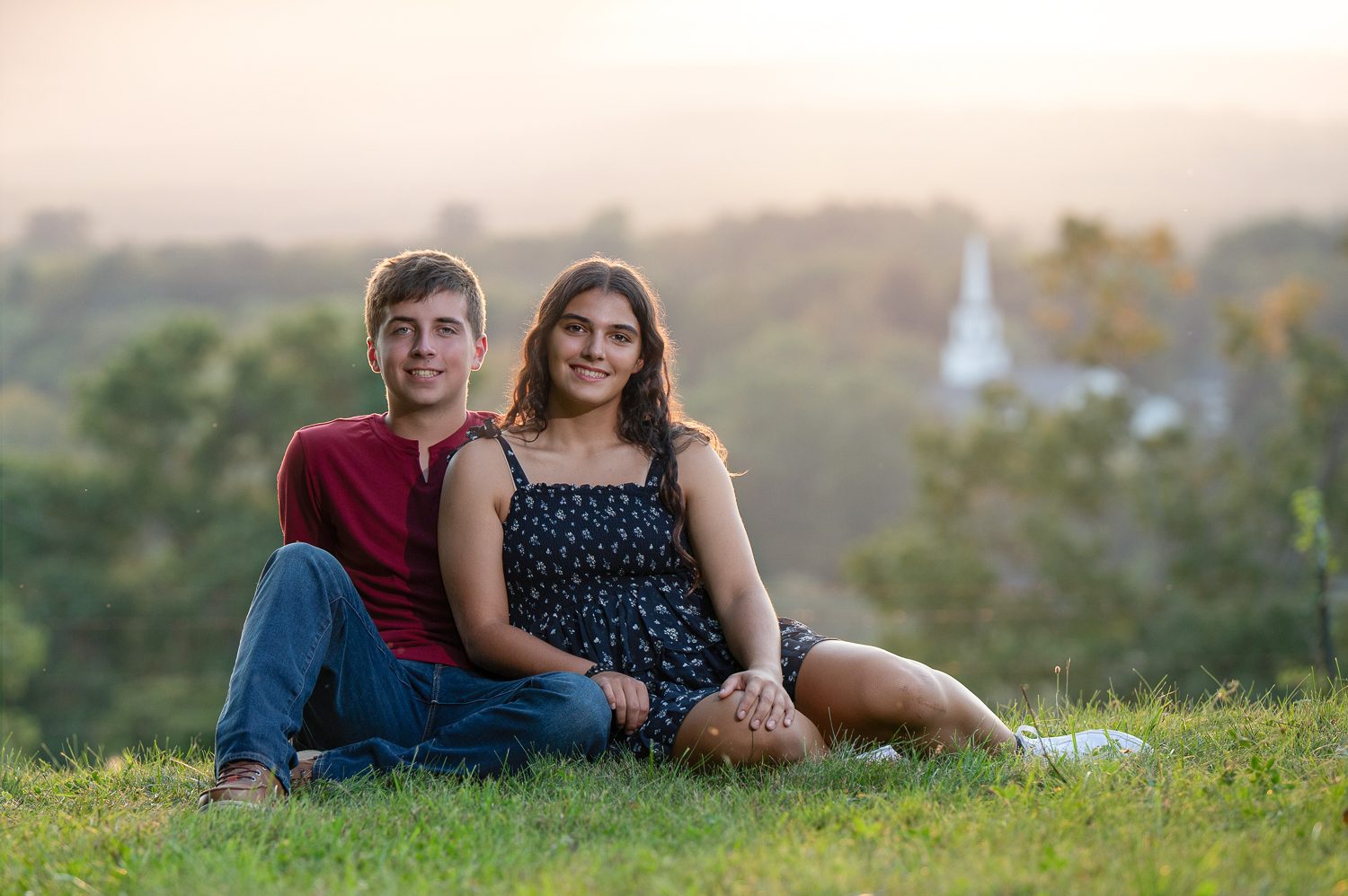 High school senior sitting on the hilltop grass with a young man in a maroon shirt and the valley behind at Gibbet Hill in Groton MA