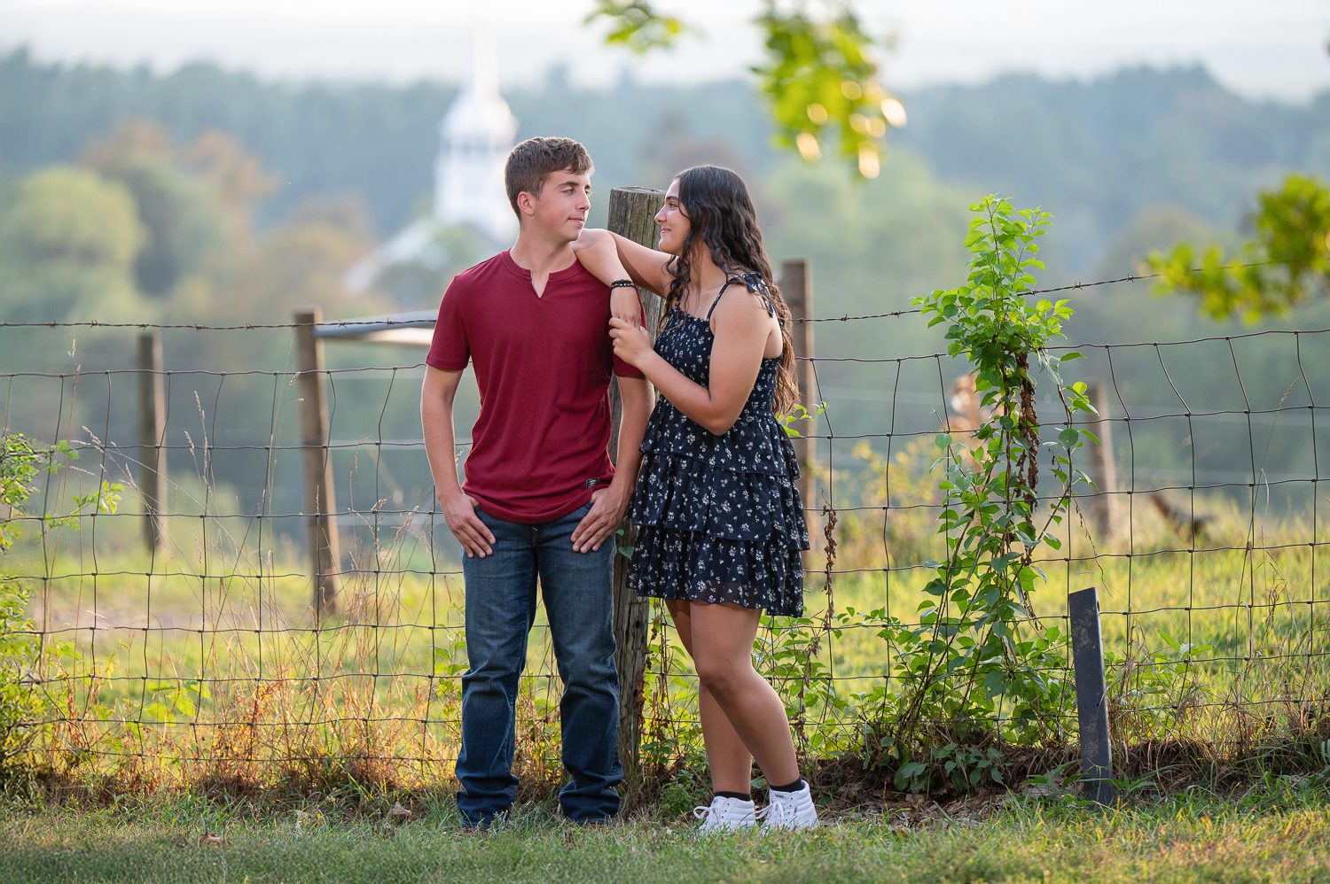 High school senior standing with a young man in a maroon shirt beside a slender tree with the Groton valley behind them