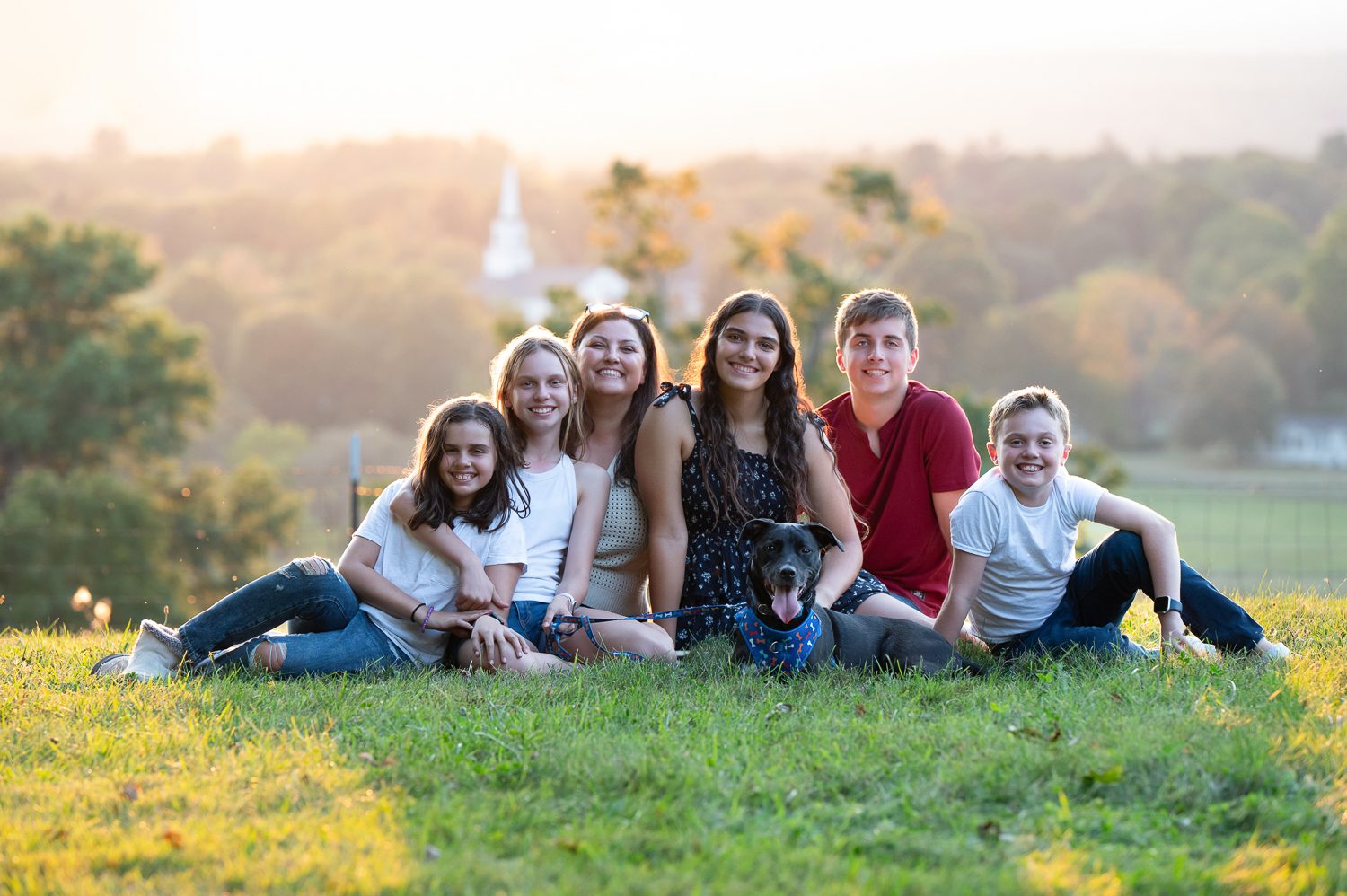 Six high school friends sitting together on the summit grass at Gibbet Hill with the valley and a church steeple behind them in Groton MA
