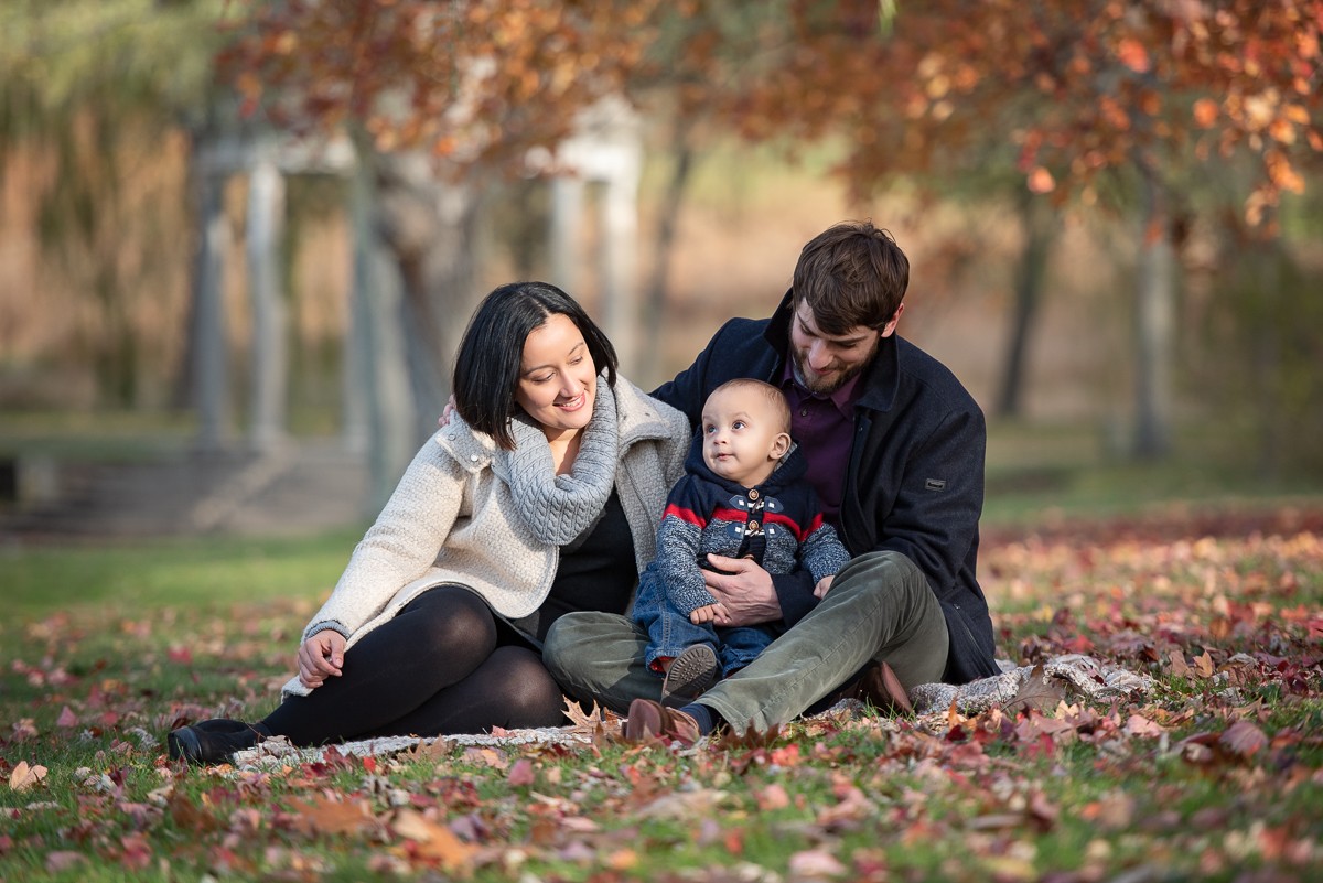 Parents sitting in a deep drift of autumn leaves with their baby son at Larz Anderson Park in Brookline Massachusetts