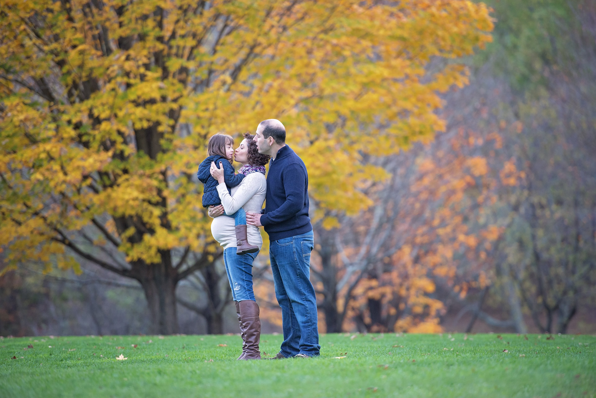 Expecting mother holding her young daughter facing her husband on the open lawn beneath a gold maple at Larz Anderson Park in Brookline Massachusetts